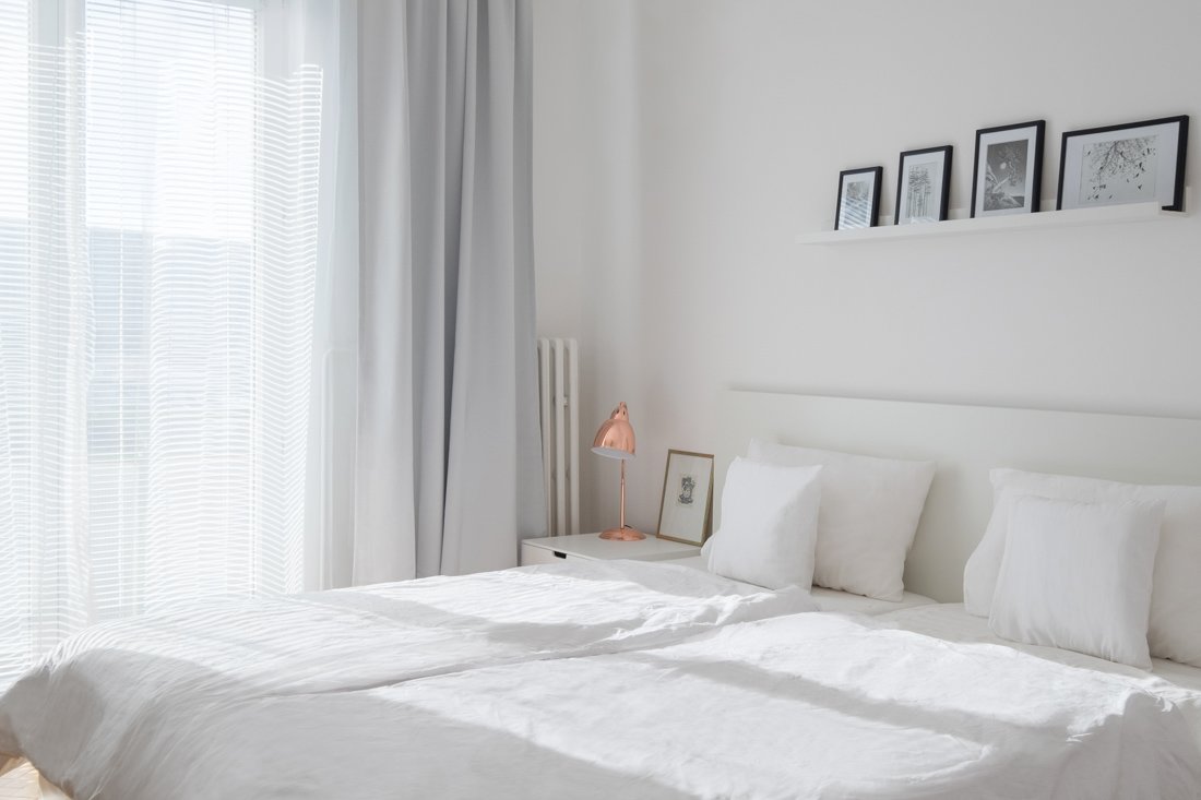 Biela spálňa s francúzskymi oknami so šedými závesmi a poličkou nad dvojposteľou