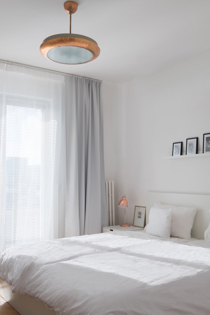 Biela spálňa v škandinávskom štýle s medenými lampičkami aj lustrom a francúzskymi oknami so šedými závesmi