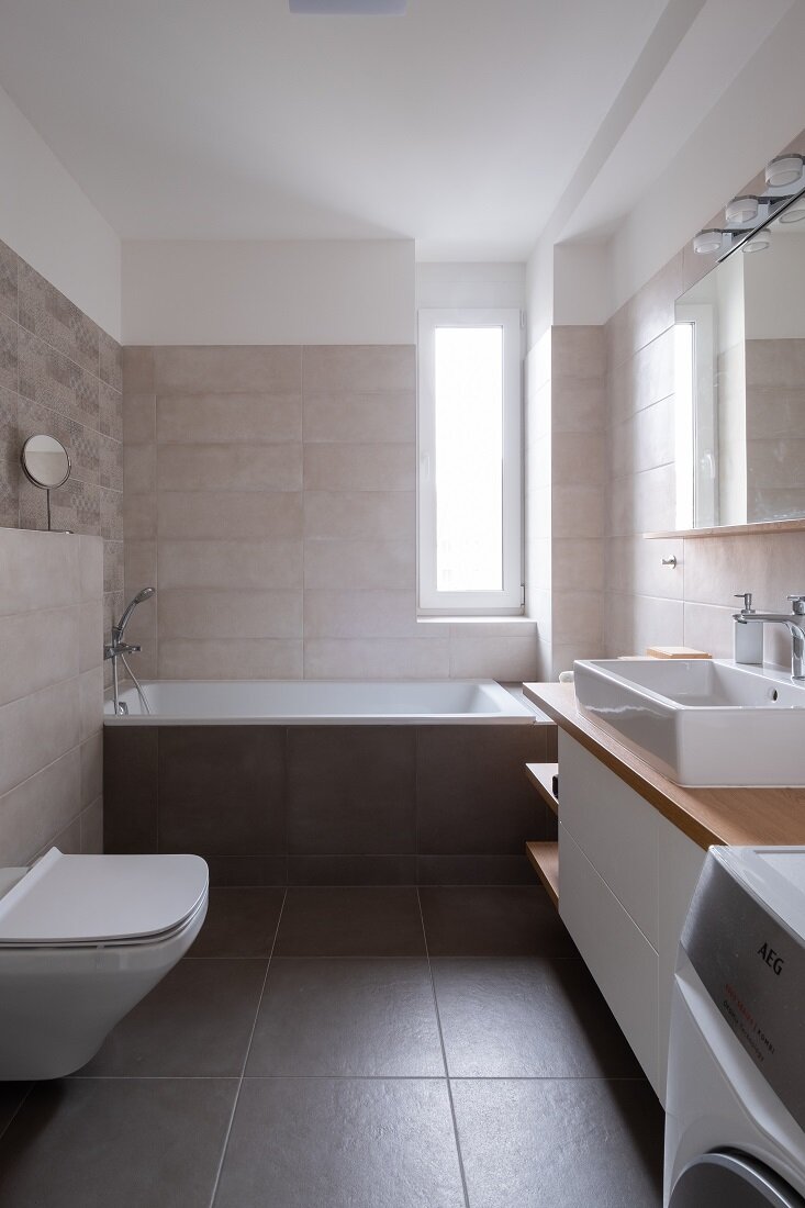 Kúpeľňa s vaňou, práčkou, záchodom, umývadlom so skrinkami na mieru a bledým obkladom v kontraste s tmavou podlahou