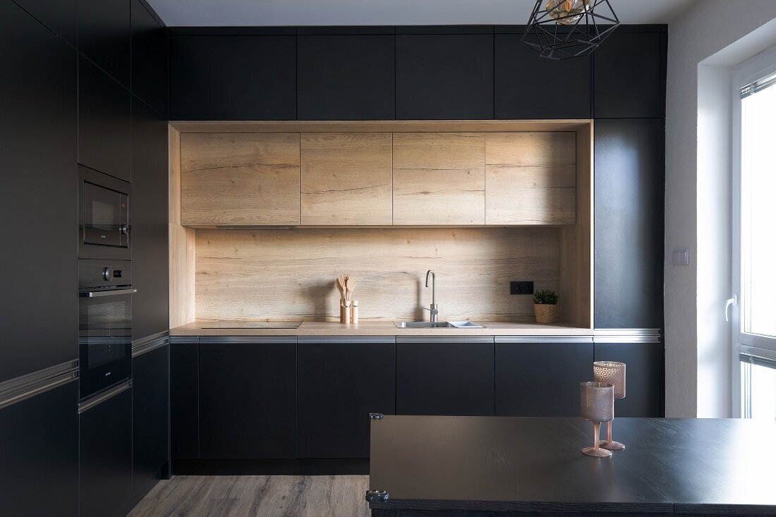 Elegantná čierna kuchyňa tvaru L s dekórom zo svetlého dreva a čiernym jedálenským stolom, osvetlená visiacim lustrom