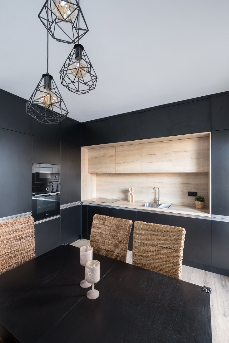 Kuchyňa kombinujúca čiernu a drevo s linkou tvaru L a štvorcovým jedálenským stolom pod visiacim kovovým lustrom