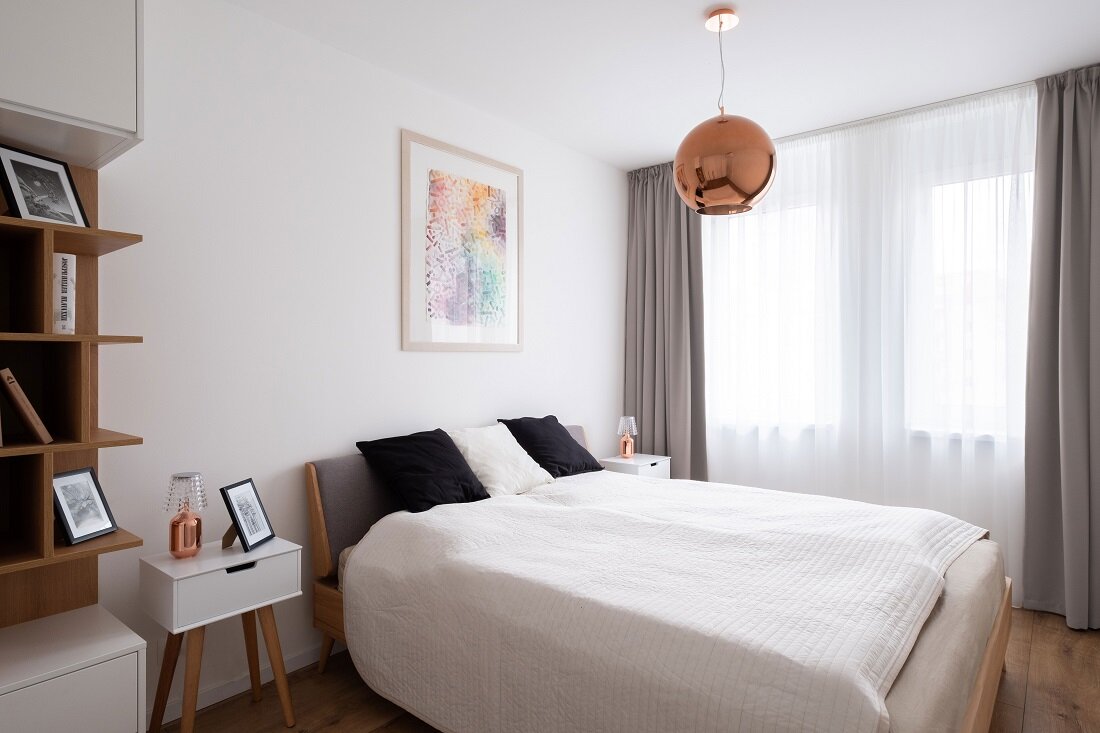 Spálňa s hlavnou farebnou kombináciou biela a drevo s prirodzeným osvetlením z okien so záclonou a závesmi
