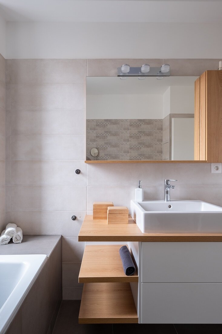 Moderná kúpeľňa s vaňou a zrkadlom na drevenej skrinke ladiacej s policami, ktoré sú časťou bielej skrinky pod umývadlom