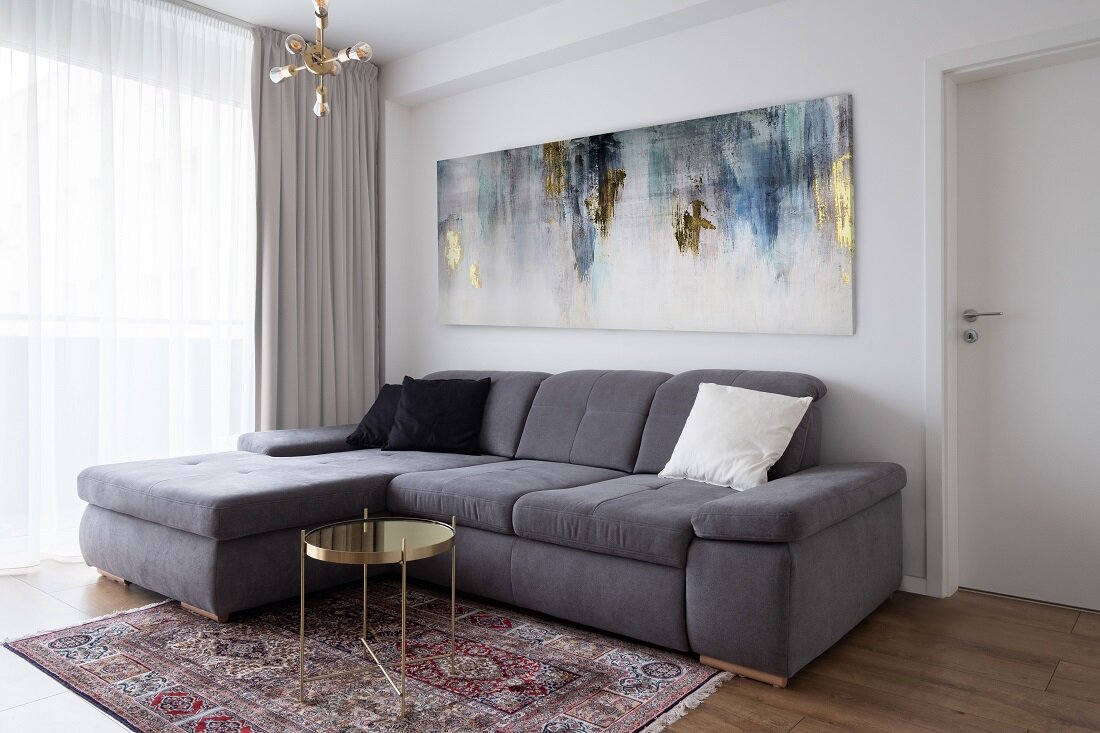 Obývačka so sivou sedačkou a malým stolíkom so zlatou farbou na vzorovanom koberci, skrášlená abstraktným obrazom