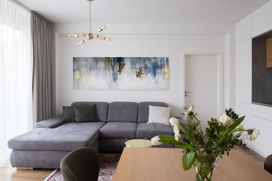 Obývačka so sivou sedačkou tvaru L a malým kruhovým stolíkom zlatej farby, zútulnená širokým abstraktným obrazom