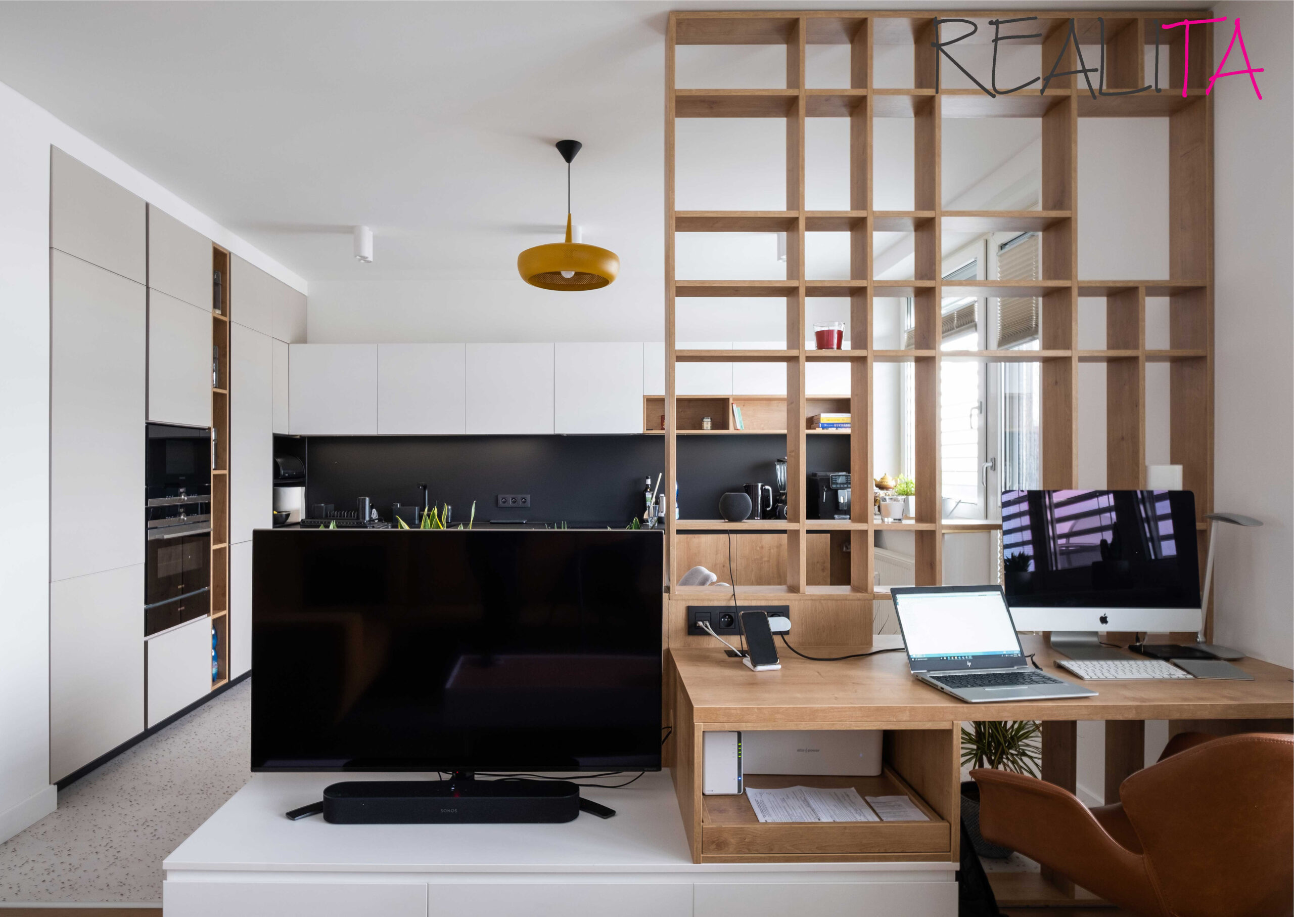 Pohľad do kuchynskej časti izby z obývačky s pracovným stolom za panelom prázdnych políc z rovnakého dreva